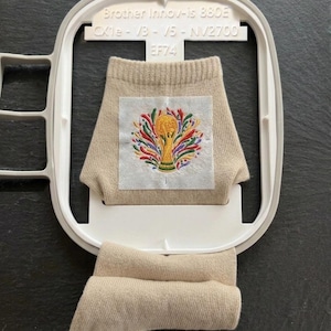 Peut inclure: Une chaussette beige fixée dans un cadre à broder blanc Brother Innov-is 880E. La chaussette présente un écusson carré avec un motif brodé coloré représentant un trophée doré entouré de feuilles stylisées rouges, vertes, bleues et violettes.