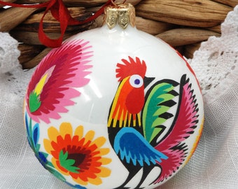 May include: White Christmas ornament with a colorful rooster and floral design. The rooster is blue, red, yellow, and green. The flowers are red, yellow, green, and pink.