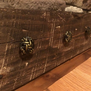 May include: A rustic wooden coat rack with three decorative metal hooks. The hooks are a dark bronze color with a woven design. The wood is a dark brown with a distressed finish.