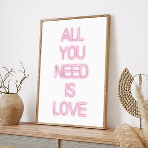 May include: A framed art print with the words "ALL YOU NEED IS LOVE" in pink neon-style lettering on a white background. The wooden frame sits on a wooden cabinet, with a vase and decorative items on either side.