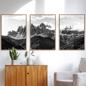May include: Three framed black and white mountain landscape prints. The artwork features dramatic peaks, clouds, and valleys. The frames are a warm brown color, and the prints are displayed above a wooden cabinet.