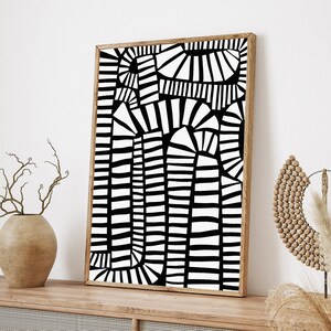 May include: A framed black and white abstract art print. The artwork features a geometric design with vertical and curved lines. The frame is a light brown colour. The print is displayed on a wooden surface.