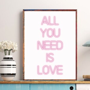 May include: A framed art print with the words "ALL YOU NEED IS LOVE" in pink neon-style lettering. The artwork is displayed on a blue cabinet, next to a vase with white flowers. A light fixture hangs from the ceiling.