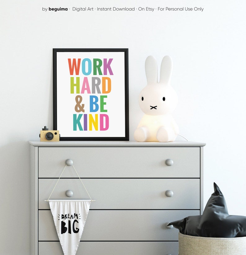May include: A framed print with the text "WORK HARD & BE KIND" in colorful letters. The print is hanging on a gray dresser with a white bunny lamp and a white pennant with the text "DREAM BIG" hanging below.