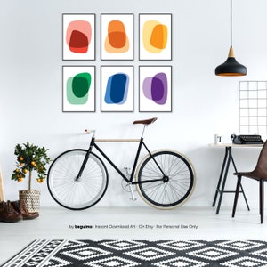 May include: Six framed abstract art prints with colorful overlapping shapes in a rainbow color scheme. The prints are hung on a white wall in a living room setting with a bicycle, a desk, and a chair.