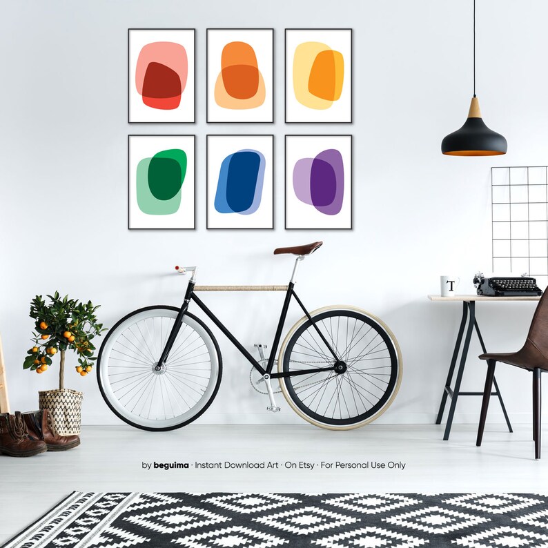 May include: Six framed abstract art prints with colorful overlapping shapes in a rainbow color scheme. The prints are hung on a white wall in a living room setting with a bicycle, a desk, and a chair.
