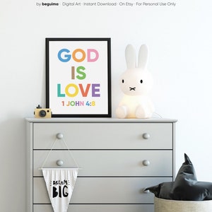May include: A framed print with the words "GOD IS LOVE" in colorful letters, with the Bible verse 1 JOHN 4:8 below. A white bunny lamp and a small wooden camera sit on a gray dresser. A banner with "DREAM BIG" hangs below.