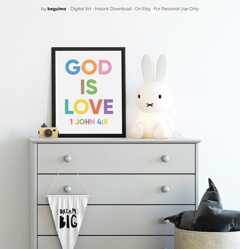 May include: A framed print with the words "GOD IS LOVE" in colorful letters, with the Bible verse 1 JOHN 4:8 below. A white bunny lamp and a small wooden camera sit on a gray dresser. A banner with "DREAM BIG" hangs below.