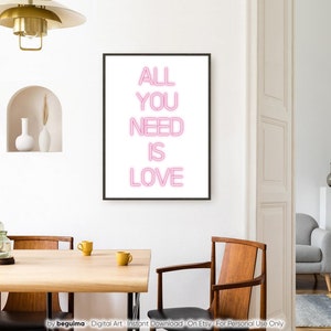 May include: A framed art print with the words "ALL YOU NEED IS LOVE" in pink neon-style lettering. The artwork is displayed in a modern dining room setting with a wooden table, chairs, and decorative elements.