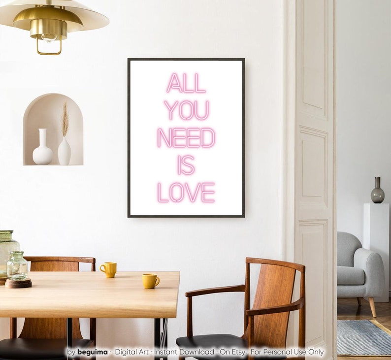 May include: A framed art print with the words "ALL YOU NEED IS LOVE" in pink neon-style lettering. The artwork is displayed in a modern dining room setting with a wooden table, chairs, and decorative elements.