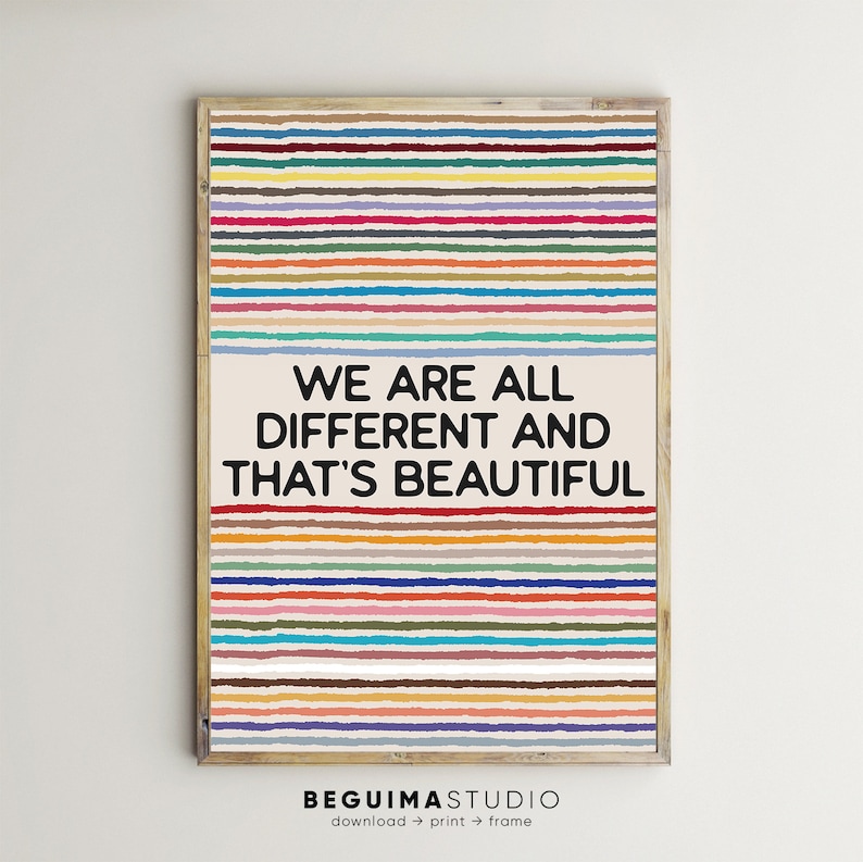 May include: A framed art print with a light wood frame. The artwork features horizontal stripes in various colours above and below a cream-coloured rectangle with the text "WE ARE ALL DIFFERENT AND THAT'S BEAUTIFUL" in black. The bottom of the print has the text "BEGUIMASTUDIO".