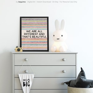 May include: A framed print with the words "WE ARE ALL DIFFERENT AND THAT'S BEAUTIFUL" above a striped background. A white bunny lamp and a small wooden camera sit on a gray chest of drawers. A banner with "DREAM BIG" hangs below.