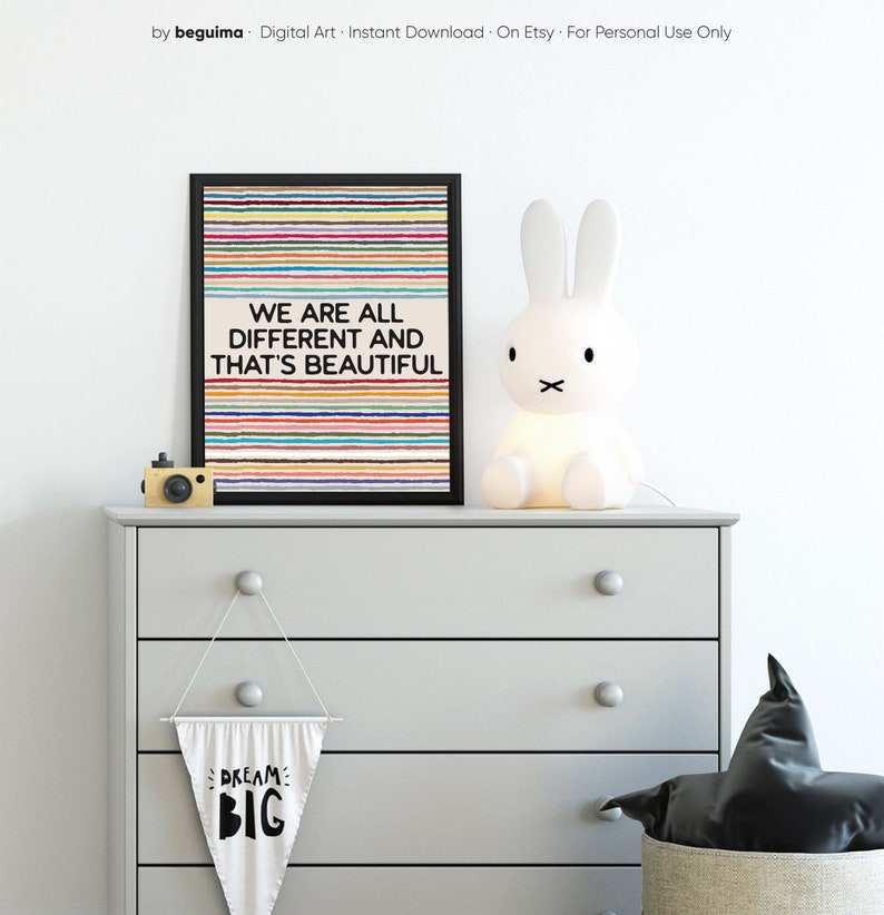 May include: A framed print with the words "WE ARE ALL DIFFERENT AND THAT'S BEAUTIFUL" above a striped background. A white bunny lamp and a small wooden camera sit on a gray chest of drawers. A banner with "DREAM BIG" hangs below.