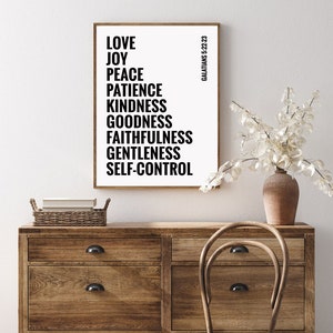 May include: A black and white printable wall art print with the text "LOVE, JOY, PEACE, PATIENCE, KINDNESS, GOODNESS, FAITHFULNESS, GENTLENESS, SELF-CONTROL" and the text "GALATIANS 5:22-23" in a smaller font. The print is framed and hanging on a white wall above a wooden dresser.