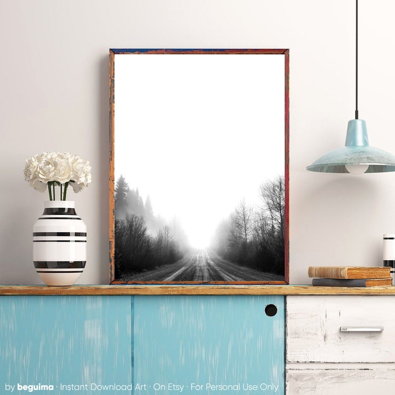 May include: Black and white framed art print of a road disappearing into a foggy forest. The print is in a rustic frame with a distressed look. A white vase with white flowers and a blue cabinet are in the foreground.