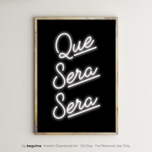Op de afbeelding: Een ingelijste kunstdruk met de woorden "Que Sera Sera" in witte neonstijl op een zwarte achtergrond. Het kunstwerk is in een houten lijst.