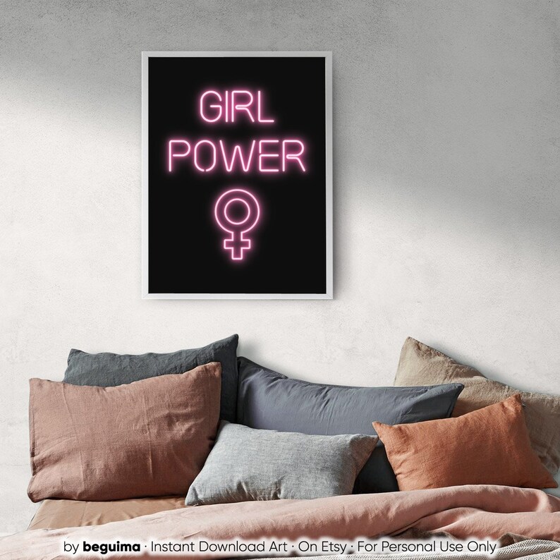 May include: A framed print featuring the words "GIRL POWER" in pink neon, and the female symbol, on a black backdrop. The artwork is set above a bed with pillows in grey, pink, and orange hues.