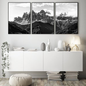 May include: A triptych of black and white landscape photographs featuring majestic mountain peaks partially obscured by clouds. The artwork is framed in black and hangs above a white cabinet with decorative vases and a lamp.