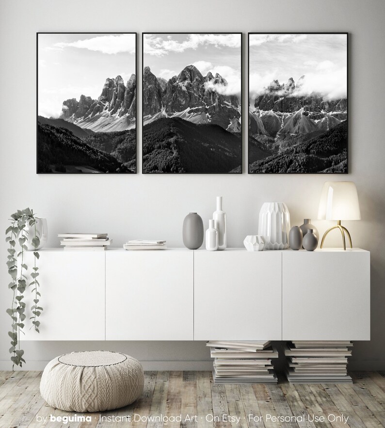 May include: A triptych of black and white landscape photographs featuring majestic mountain peaks partially obscured by clouds. The artwork is framed in black and hangs above a white cabinet with decorative vases and a lamp.