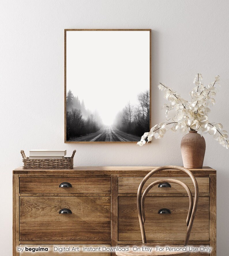 May include: Black and white framed art print of a road disappearing into a foggy forest. The artwork is displayed above a wooden dresser with a woven basket, books, and a vase with white flowers.
