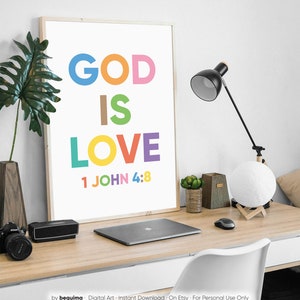 May include: A framed print with the words "GOD IS LOVE" in colorful block letters, with "1 JOHN 4:8" below. The print is on a desk with a laptop, lamp, and decorative items. The overall theme is inspirational and religious.