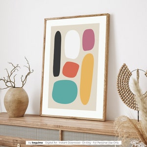 May include: A framed abstract art print featuring colorful, organic shapes in black, white, red-orange, pink, yellow, and teal on a beige background. The artwork is displayed in a wooden frame and rests on a wooden surface.