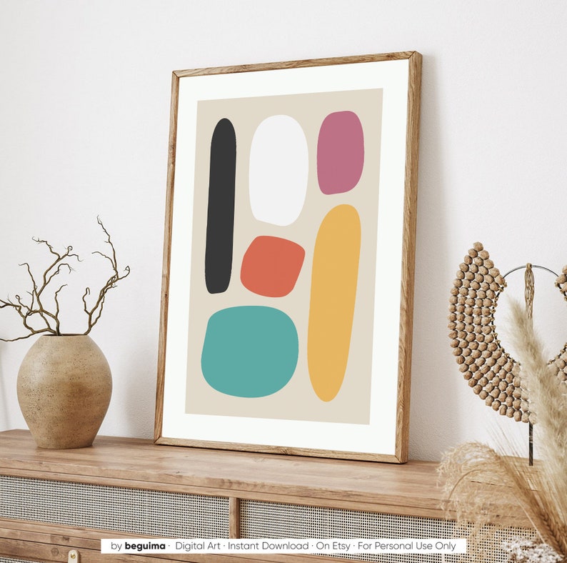 May include: A framed abstract art print featuring colorful, organic shapes in black, white, red-orange, pink, yellow, and teal on a beige background. The artwork is displayed in a wooden frame and rests on a wooden surface.