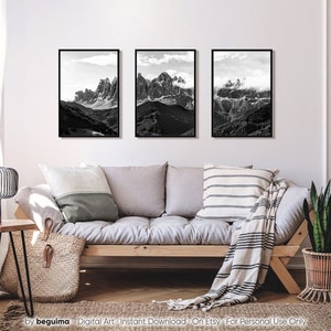 May include: Three framed black and white mountain landscape prints. The images depict rugged peaks and valleys under cloudy skies. The prints are displayed above a light-colored sofa with neutral-toned pillows and a woven throw.