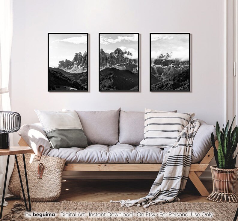 May include: Three framed black and white mountain landscape prints. The images depict rugged peaks and valleys under cloudy skies. The prints are displayed above a light-colored sofa with neutral-toned pillows and a woven throw.