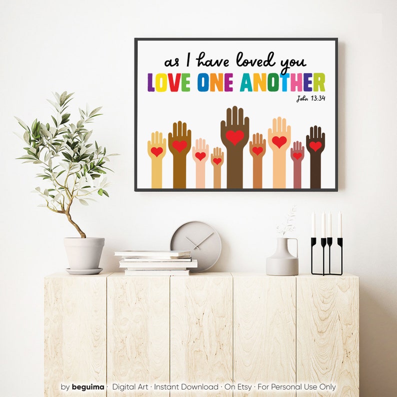 May include: A digital art print with a white background and a black frame. The print features a quote from the Bible, "as I have loved you LOVE ONE ANOTHER John 13:34". The quote is in rainbow colors. Below the quote are eight hands of different skin tones, each with a red heart in the palm of the hand.