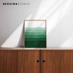 May include: A framed art print featuring a watercolor design of layered green hues. The artwork has a gold frame and is displayed on a wooden cabinet. The top of the print has a white background, with the text "BEGUIMA STUDIO" at the top.