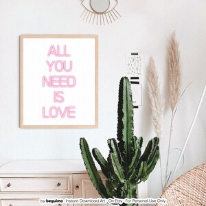 May include: A framed print with the words "ALL YOU NEED IS LOVE" in pink neon-style lettering. The artwork is displayed on a white wall, alongside a cactus, pampas grass, and a decorative eye-shaped mirror.