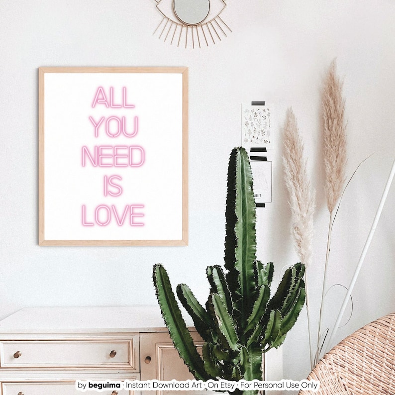 May include: A framed print with the words "ALL YOU NEED IS LOVE" in pink neon-style lettering. The artwork is displayed on a white wall, alongside a cactus, pampas grass, and a decorative eye-shaped mirror.