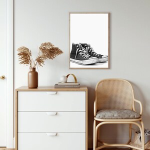 Puede incluir: Impresi&oacute;n en blanco y negro enmarcada con un par de zapatillas de ca&ntilde;a alta. Las zapatillas son negras con cordones blancos y suela blanca. La impresi&oacute;n se muestra en una pared blanca sobre una c&oacute;moda blanca con un jarr&oacute;n marr&oacute;n y plantas secas.