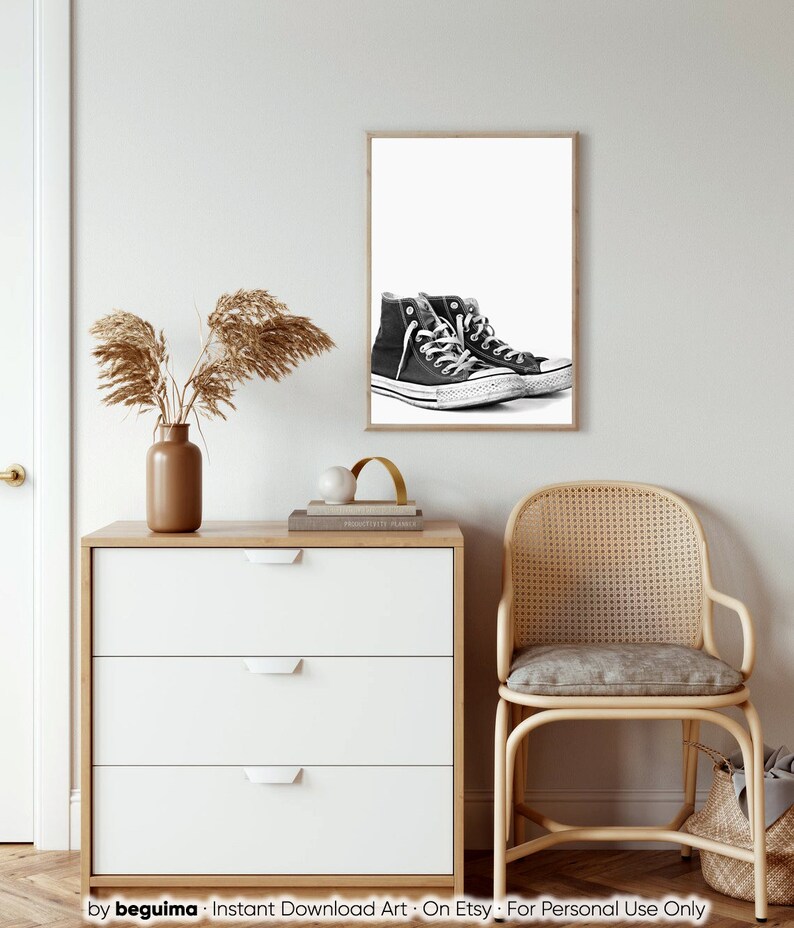 Puede incluir: Impresi&oacute;n en blanco y negro enmarcada con un par de zapatillas de ca&ntilde;a alta. Las zapatillas son negras con cordones blancos y suela blanca. La impresi&oacute;n se muestra en una pared blanca sobre una c&oacute;moda blanca con un jarr&oacute;n marr&oacute;n y plantas secas.