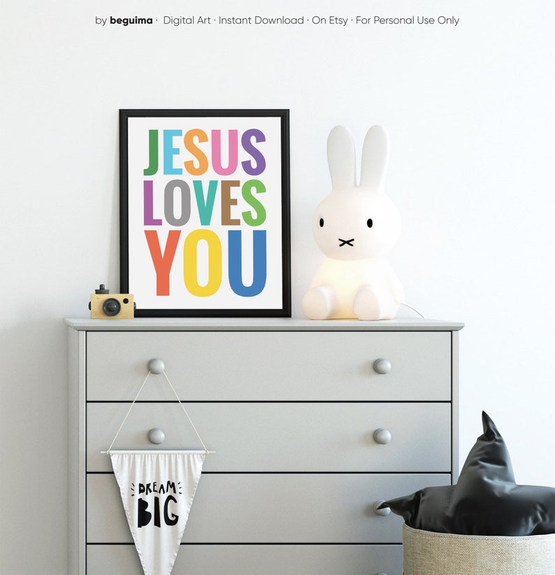 Może przedstawiać: Wydruk w ramie z tekstem "JESUS LOVES YOU" w kolorach tęczy. Wydruk wisi na białej ścianie nad komodą z białą lampą w kształcie kr&oacute;lika i czarno-białym aparatem.