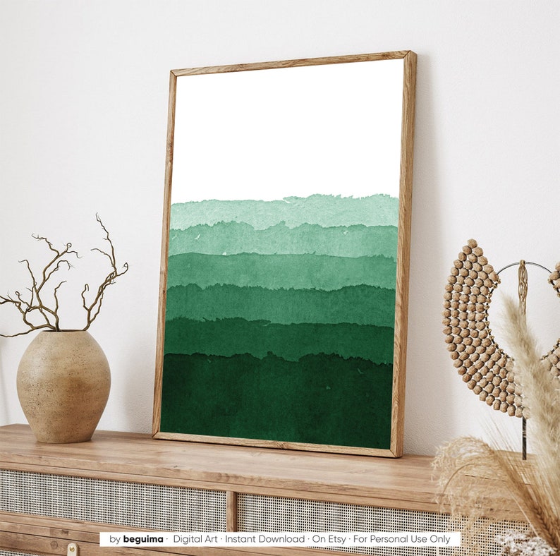 May include: A framed art print featuring an abstract watercolor design in shades of green. The artwork has a white upper section and layered bands of varying green hues. The wooden frame complements the artwork's natural aesthetic. The print is displayed on a wooden surface.