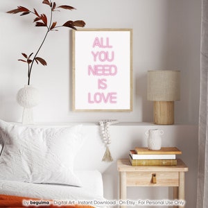 May include: A framed print with the words "ALL YOU NEED IS LOVE" in pink neon-style lettering. The artwork is displayed on a white wall, alongside a lamp, books, and decorative items. The overall aesthetic is modern and minimalist.