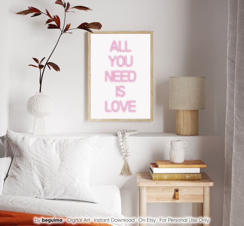 May include: A framed print with the words "ALL YOU NEED IS LOVE" in pink neon-style lettering. The artwork is displayed on a white wall, alongside a lamp, books, and decorative items. The overall aesthetic is modern and minimalist.
