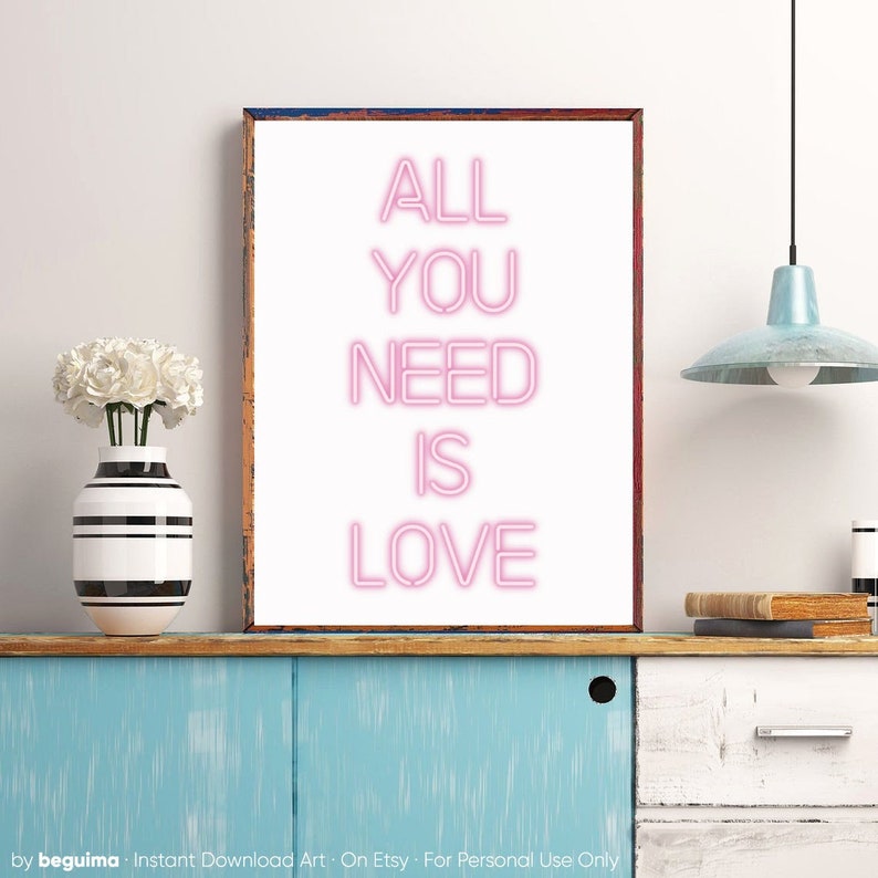May include: A framed art print with the words "ALL YOU NEED IS LOVE" in pink neon-style lettering. The artwork is displayed on a blue cabinet, next to a vase with white flowers. A light fixture hangs from the ceiling.