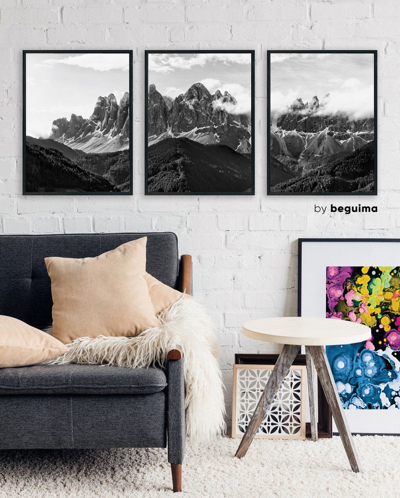 May include: Three framed black and white mountain landscape prints hang on a white brick wall. The artwork depicts rugged peaks with snow and clouds. The prints are above a gray sofa with beige pillows and a white fur throw.