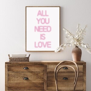 May include: A framed print with the words "ALL YOU NEED IS LOVE" in pink neon-style lettering. The artwork is displayed on a wooden dresser with a vase of white flowers and a wicker basket.
