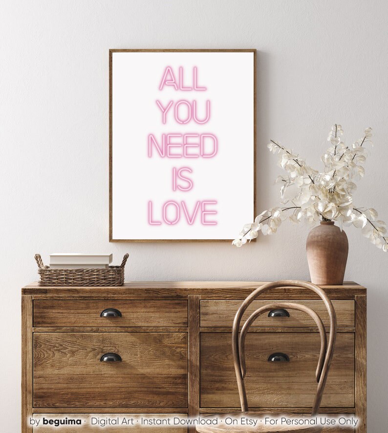 May include: A framed print with the words "ALL YOU NEED IS LOVE" in pink neon-style lettering. The artwork is displayed on a wooden dresser with a vase of white flowers and a wicker basket.