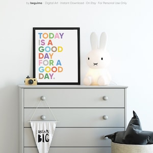 May include: A white dresser with four drawers and a black and white pennant hanging from the second drawer that says "Dream Big". A framed print with the text "Today is a good day for a good day." in colorful letters is on top of the dresser. A white bunny lamp is on the left side of the dresser.
