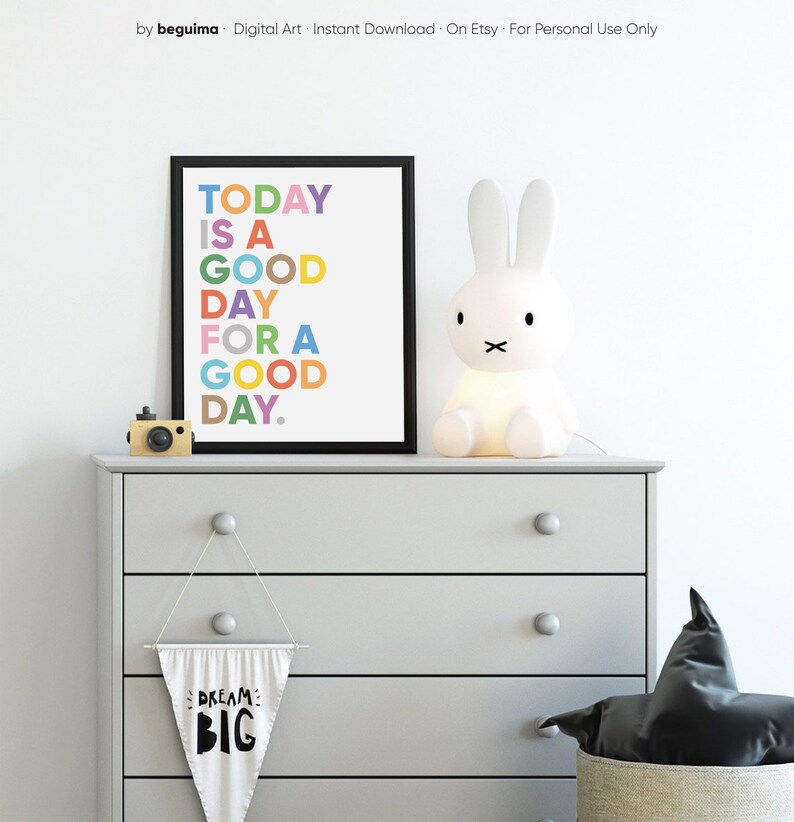 May include: A white dresser with four drawers and a black and white pennant hanging from the second drawer that says "Dream Big". A framed print with the text "Today is a good day for a good day." in colorful letters is on top of the dresser. A white bunny lamp is on the left side of the dresser.