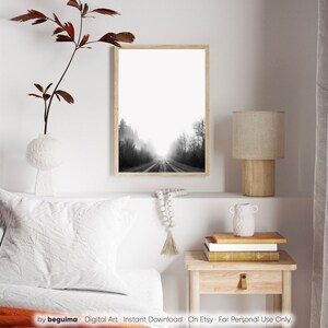 May include: A framed black and white photograph of a road disappearing into a misty forest. The artwork is displayed on a white wall, alongside a decorative vase with a branch, a lamp, and a small wooden side table. The image is a digital art print.