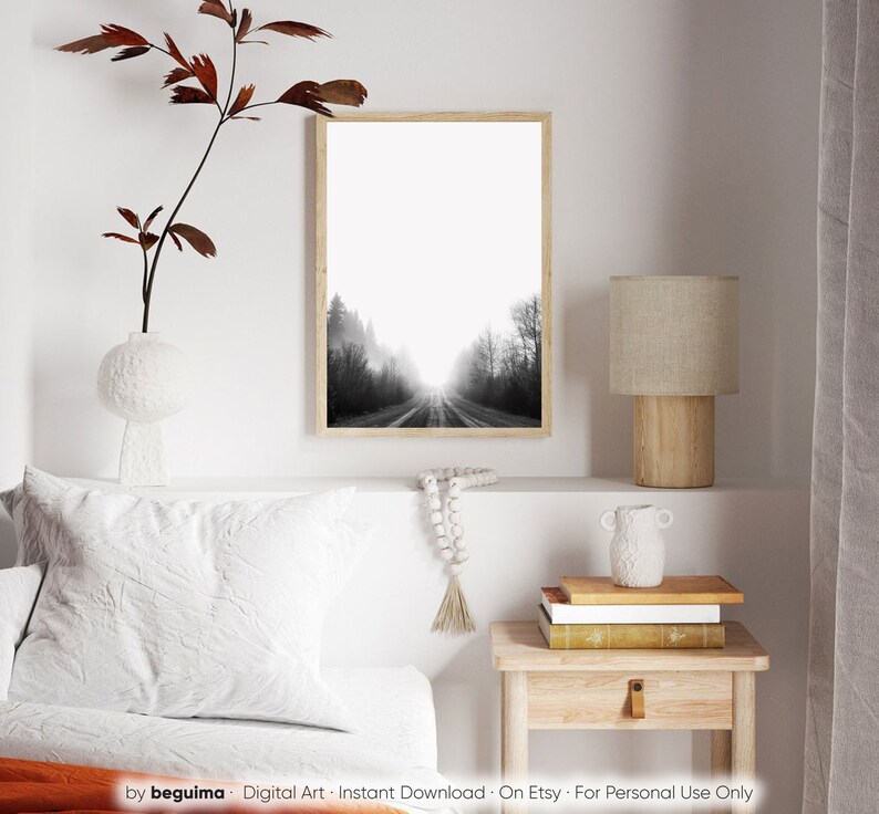 May include: A framed black and white photograph of a road disappearing into a misty forest. The artwork is displayed on a white wall, alongside a decorative vase with a branch, a lamp, and a small wooden side table. The image is a digital art print.