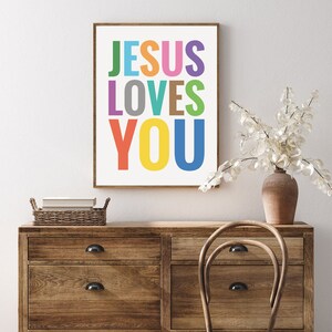 Może przedstawiać: Kolorowy, drukowany obraz na ścianę z tekstem "JESUS LOVES YOU" w tęczowych kolorach. Obraz jest oprawiony i wisi na ścianie nad drewnianym komodą.