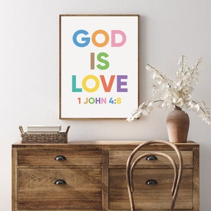 May include: A framed art print with the words "GOD IS LOVE" in colorful letters. Below the words is the text "1 JOHN 4:8". The print is displayed on a wooden dresser with a vase of white flowers.