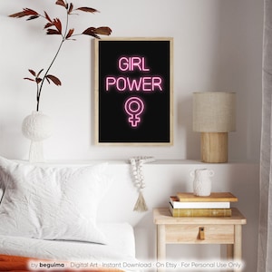 May include: A framed art print featuring the words "GIRL POWER" in vibrant pink neon, and the female symbol, set against a black backdrop. The print is in a light wood frame. The artwork is displayed on a white shelf with decorative items.